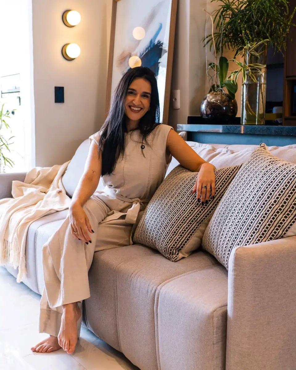 Larissa Cardin, arquiteta, sorrindo sentada no sofá de um apartamento entregue, com arandelas acesas, quadro e plantas ao fundo